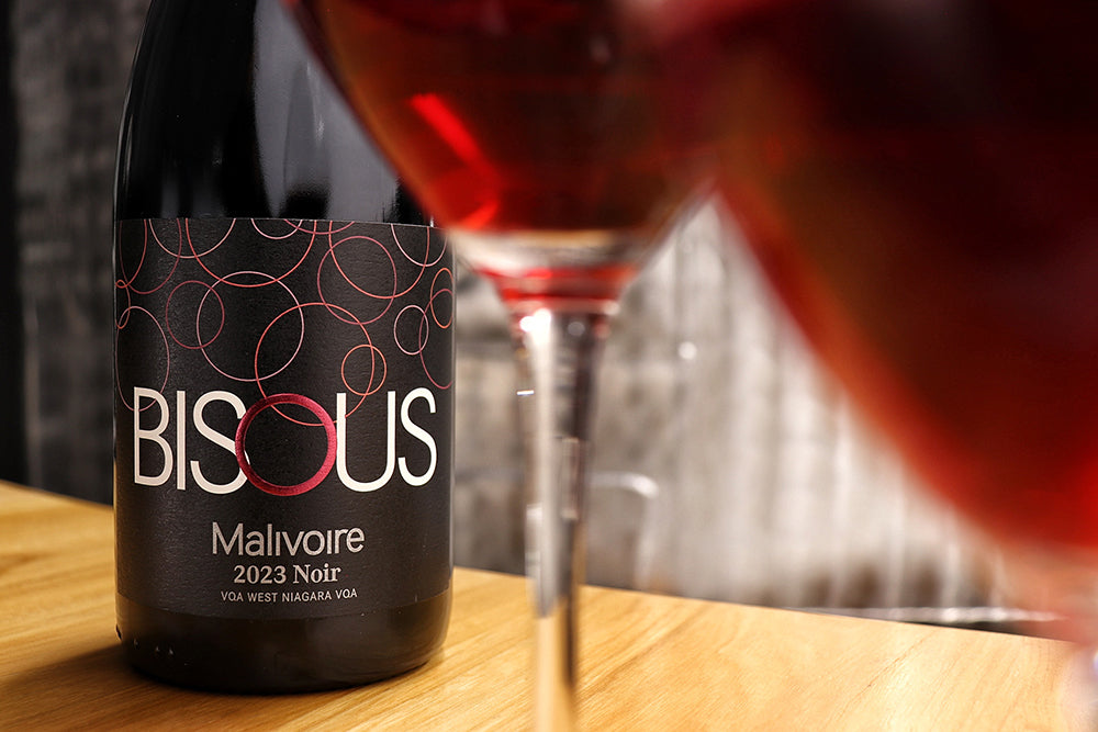 Bottle of Bisous Malivoire 2023 Noir wine with a glass of red wine on a wooden surface.